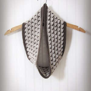 Puede incluir: Una bufanda infinita de crochet marrón y beige colgada de una percha de madera. La bufanda está hecha con un patrón de punto texturizado.