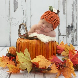 May include: A newborn baby wearing a crocheted pumpkin hat sleeps in a pumpkin-shaped basket surrounded by fall leaves.