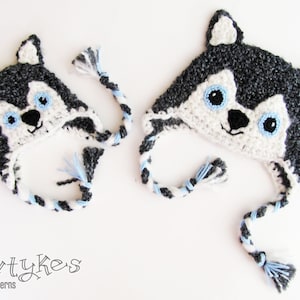 Könnte beinhalten: Zwei gehäkelte Husky-Mützen mit blauen und weißen Ohrenklappen und -Schwanz. Die Mützen sind grau und weiß mit schwarzen Nasen und blauen Augen.