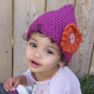 May include: A young child wearing a pink crocheted hat with a large orange crocheted flower on the side. The hat has a pointed top and a textured design.
