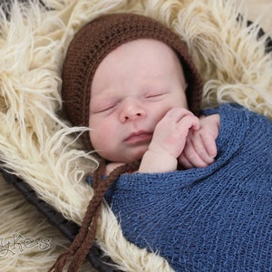 May include: A newborn baby wearing a brown crocheted hat and a blue knitted wrap, sleeping in a basket lined with white faux fur.