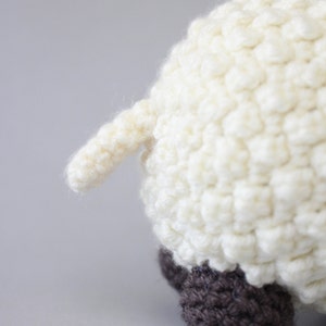 May include: A white crocheted sheep with brown crocheted feet. The sheep is facing away from the camera and its tail is visible.