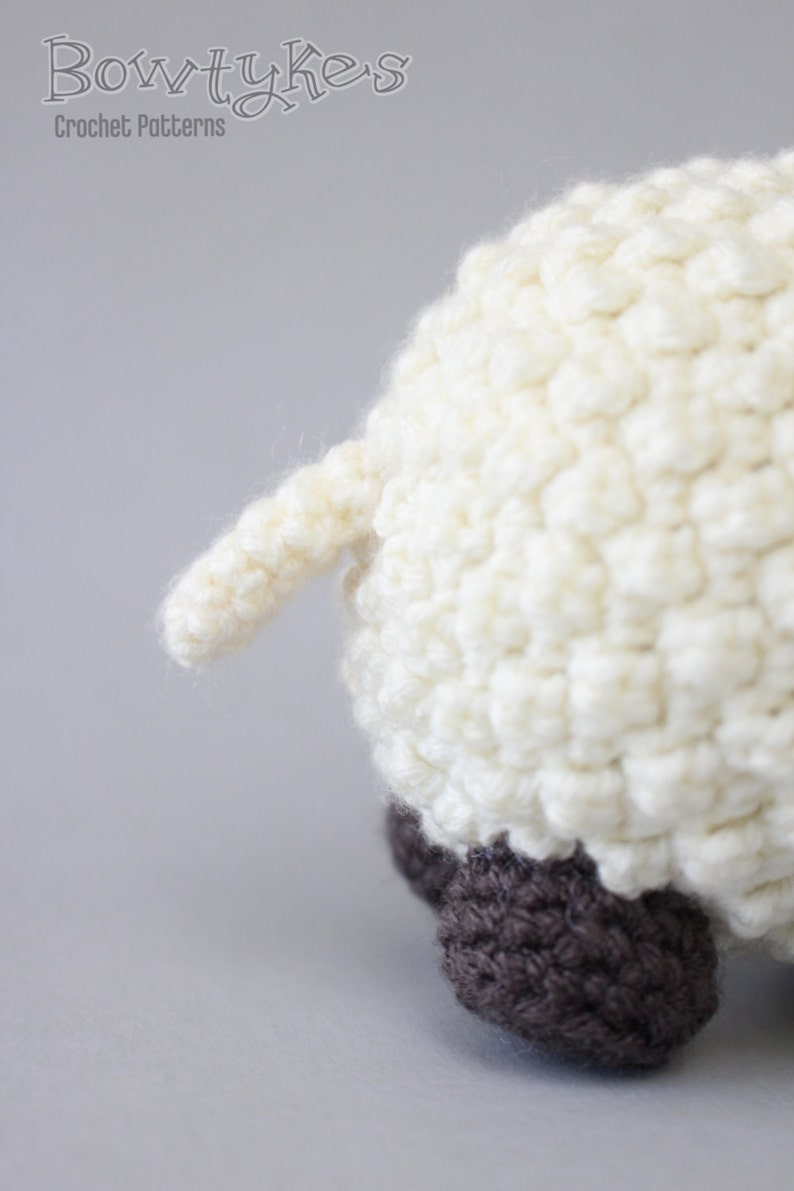 May include: A white crocheted sheep with brown crocheted feet. The sheep is facing away from the camera and its tail is visible.