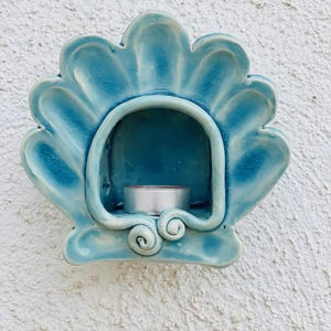 May include: A wall-mounted ceramic candle holder shaped like a scalloped seashell. The piece features a glossy, crackled glaze in shades of light and dark blue. A small, round, silver-colored metal tea light candle sits inside the central cavity.