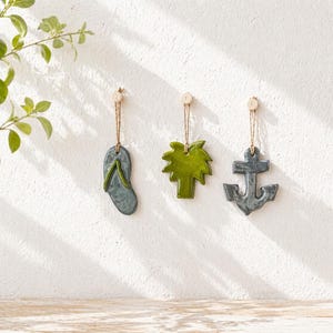 Op de afbeelding: Drie keramische wandornamenten: een slipper, een palmboom en een anker. De slipper is grijs met een groene band. De palmboom is groen en het anker is grijs. Elk ornament hangt aan een houten peg met touw.