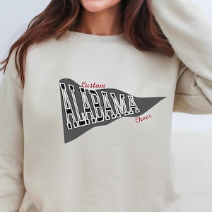 Puede incluir: Una sudadera de cuello redondo y manga larga en color beige con un gráfico de banderín gris oscuro en el pecho. El banderín contiene el texto blanco ALABAMA con bordes negros, con el texto rojo en cursiva Custom arriba y Cheer abajo.