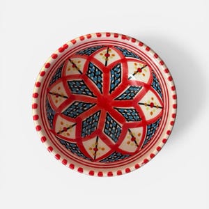 Może przedstawiać: Ozdobna ceramiczna miska z żywym kwiatowym wzorem. Miska ma czerwono-białą krawędź z serią czerwonych kropek. Wnętrze prezentuje symetryczny wzór z czerwonymi, niebieskimi i kremowymi elementami. Miska jest prawdopodobnie używana do serwowania jedzenia lub jako element dekoracyjny.