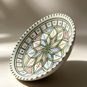 Puede incluir: Plato ovalado de cerámica con un diseño floral colorido. El plato presenta un borde verde con pequeños puntos y un patrón central de formas verdes, azules, amarillas y rojas. El plato está ligeramente inclinado.