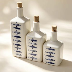 Puede incluir: Tres botellas de cerámica blanca de diferentes alturas, cada una con base cuadrada y tapón de corcho. El frente de cada botella está decorado con un patrón vertical de motivos de peces pintados en azul separados por pequeños puntos azules.