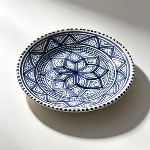 Puede incluir: Un plato redondo de cerámica con un detallado patrón geométrico azul y blanco. El plato presenta un diseño floral central rodeado de formas intrincadas y bordes punteados. La paleta de colores incluye tonos de azul y blanco.