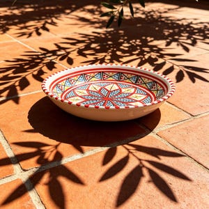 May include: A decorative ceramic bowl with a vibrant, geometric pattern in red, blue, yellow, and white. The bowl has a white base and a red rim with a dotted design. The bowl is sitting on a terracotta tile surface.