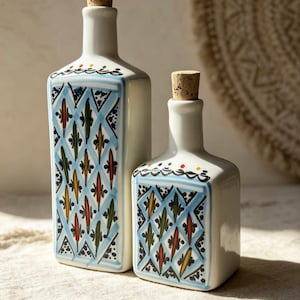 Puede incluir: Dos botellas de cerámica blanca de diferentes alturas, cada una con un tapón de corcho. Las botellas presentan un patrón de rombos azules pintado a mano, relleno con pequeñas formas de hojas coloridas en rojo, amarillo y verde con bordes azules.