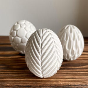 May include: Three white decorative eggs with different textured surfaces. One egg has a leaf-like pattern, another has a bubble design, and the third has a teardrop pattern. The eggs are displayed on a wooden surface.
