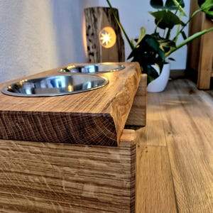Puede incluir: Una estación de alimentación elevada para mascotas hecha de madera natural con una superficie superior de borde vivo. El soporte cuenta con dos aberturas circulares que sostienen cuencos de acero inoxidable extraíbles con veta visible.