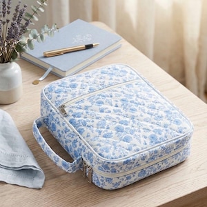Op de afbeelding: Een rechthoekig, gewatteerd stoffen etui met een blauw en wit bloemenpatroon. Het etui is voorzien van een ritssluiting, een handvat aan de zijkant en een voorvak met rits. De buitenkant is gemaakt van zacht, gewatteerd materiaal.