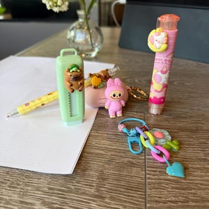 May include: A collection of colorful stationery and accessories on a wooden surface. Items include a light green eraser, a pink eraser with the word "NICOR" printed on it, a yellow pencil, and a keychain with a heart charm.