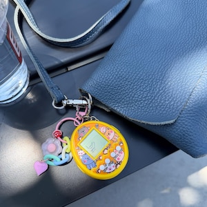 May include: A yellow digital pet toy with a screen displaying pixelated graphics and a colorful charm keychain attached to a blue leather crossbody bag. The toy features cartoon animal illustrations and three buttons labeled A, B, and C.