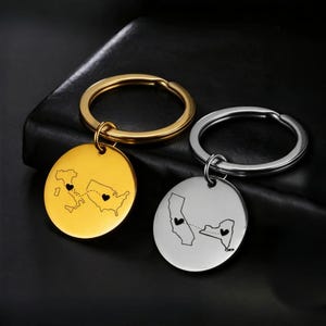 May include: Two keyrings with circular pendants. One is gold-coloured, the other silver. Each pendant shows a map outline with a heart marking two locations, connected by a dashed line, symbolising a long-distance relationship.