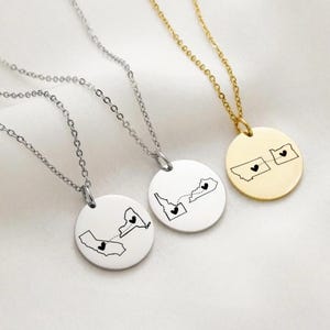 Puede incluir: Tres collares con colgantes redondos. Cada colgante presenta el contorno de un mapa de dos estados conectados por una línea de puntos y un corazón. Un collar es dorado, los otros dos son plateados.
