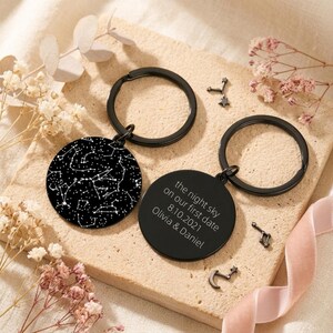 Peut inclure: Deux porte-clés ronds noirs. L'un présente un motif de constellation blanc sur fond noir. L'autre porte-clés porte l'inscription "the night sky on our first date 8.10.2021 Olivia & Daniel". Des breloques étoiles décoratives sont éparpillées.