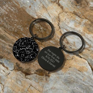 Puede incluir: Dos llaveros negros con colgantes redondos. Un colgante muestra un patrón de constelación blanco sobre un fondo negro. El otro colgante tiene el texto "the night sky on our first date 8.10.2021 Olivia & Daniel."