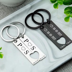 Op de afbeelding: Twee bijpassende sleutelhangers, één zilverkleurig en één zwart, elk met een rechthoekig label. De zilveren sleutelhanger heeft de letters "P", "O" en "S" met een hartsymbool. De zwarte sleutelhanger heeft de datum "06-09-2022" en de initialen "Y&K".