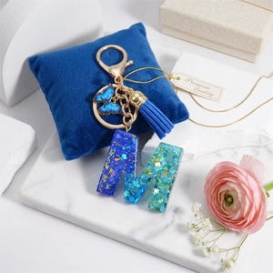 May include: A gold-toned keychain with a blue tassel and butterfly charm, and two letter "M" pendants. The pendants are filled with blue and green glitter. The keychain rests on a blue velvet pillow.
