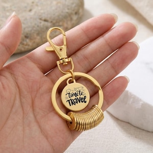 Puede incluir: Un llavero de metal dorado con cierre de mosquetón, una anilla circular y un pequeño dije redondo grabado con el texto Time to Travel, acompañado de iconos de un avión y nubes, además de una serie de anillos metálicos cilíndricos apilados.