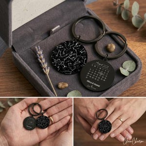 May include: Black metal keyrings with circular pendants. One pendant shows a star map, the other a calendar for August 2021, with a date highlighted. The keyrings are displayed in a grey velvet box, with a sprig of lavender.
