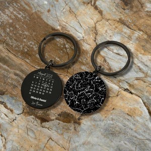 May include: Two black keychains. One keychain features a calendar for August 2021 with the names Olivia & Daniel and the words "Love Forever". The other keychain displays a white constellation pattern on a black background.