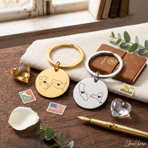 Puede incluir: Dos llaveros redondos con mapas y diseños de corazón, uno dorado y otro plateado, junto a un pequeño cuaderno, sellos y corazones decorativos. El llavero dorado muestra Italia y EE. UU. El llavero plateado muestra California y Nueva York.