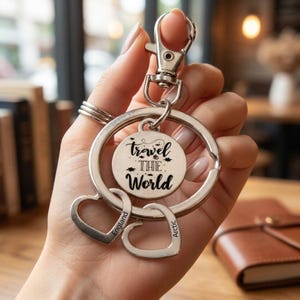 Puede incluir: Un llavero de metal plateado con un dije circular grabado con el texto travel THE World, acompañado de dos dijes en forma de corazón. Un corazón está grabado con la palabra England y el otro con la palabra Arctic, todo unido a una argolla.