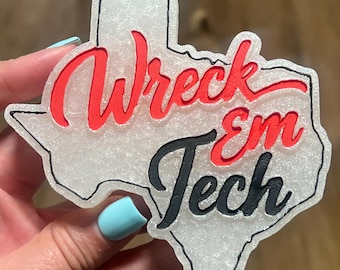 May include: A white glittery Texas-shaped ornament featuring the red text Wreck and the black text Em Tech in a cursive font. The state outline is defined by a thin black border, creating a distinct shape with a textured, sparkling surface finish.