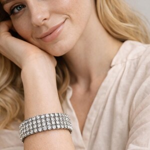 May include: A sparkling, silver-toned bracelet with multiple rows of square-cut, clear crystals. The bracelet is worn on a wrist, and the image is of a person with blonde hair and a light-colored blouse.