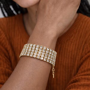 May include: A gold-toned bracelet with multiple rows of sparkling, clear crystals. The bracelet is worn on a wrist, and the clasp is visible. The background is a solid, warm orange color.