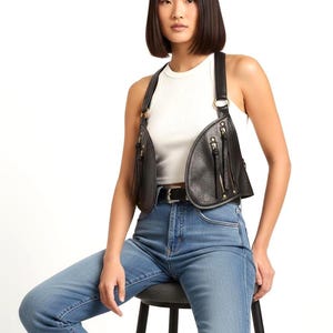 May include: A black leather vest with a unique design, featuring two front pockets with zippers and gold-colored hardware. The vest has adjustable straps and is worn over a white tank top and blue jeans.