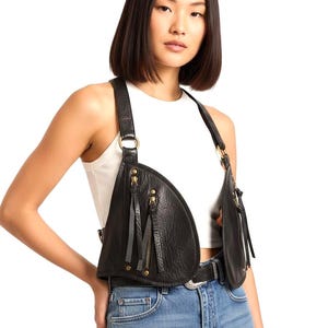 May include: Black leather harness with two asymmetrical pockets, each with a zipper and tassel detail. The harness has adjustable straps and gold-colored hardware. The model is wearing the harness over a white top and blue jeans.