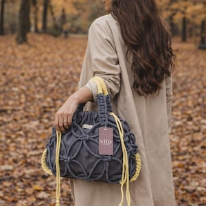 Peut inclure: Un sac à main macramé gris avec des accents jaunes et une étiquette "vita". Le sac présente un motif tissé et une fermeture à cordon. Les poignées sont jaunes. La personne porte un manteau beige.