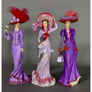 May include: Three Victorian-era figurines of women in long dresses and elaborate hats. The figurines are in shades of purple, with red and white accents. Each woman holds accessories like a purse or flowers.