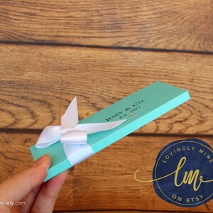 May include: A rectangular, light turquoise gift box featuring a white ribbon bow tied around the center. The box is printed with the black text "BABY & CO. Est. 2021" and includes a small, clear, circular embellishment near the printed text.