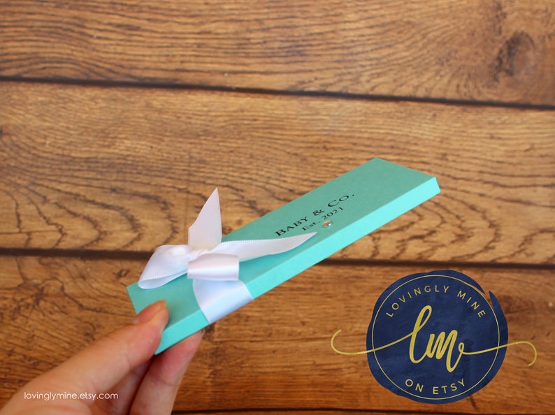 May include: A rectangular, light turquoise gift box featuring a white ribbon bow tied around the center. The box is printed with the black text "BABY & CO. Est. 2021" and includes a small, clear, circular embellishment near the printed text.