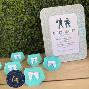 May include: A silver glitter frame with a white card inside that says "The Dirty Diaper Game". The card has a black silhouette of a man and woman with a baby. There are 7 teal blue diaper-shaped paper cutouts with white bows on a wooden surface.