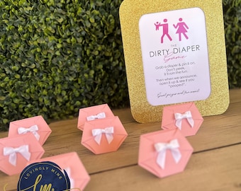 Peut inclure: Un cadre photo doré à paillettes avec un fond blanc et du texte rose qui dit "The Dirty Diaper Game". Le texte explique les règles du jeu. Il y a des découpes de papier en forme de couches roses et blanches sur une surface en bois.