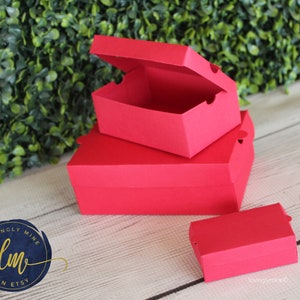May include: Three red cardboard boxes of different sizes. The largest box is open and the smallest box is closed. The boxes are on a white wooden surface.