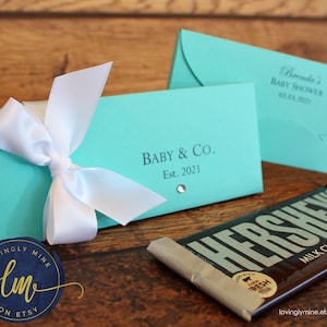 May include: Two rectangular turquoise paper candy bar wrappers with white satin ribbons. One wrapper features the text Baby & Co. Est. 2021 with a small rhinestone, and the other displays Brenda's Baby Shower 03.03.2021. A Hershey's milk chocolate bar is visible.