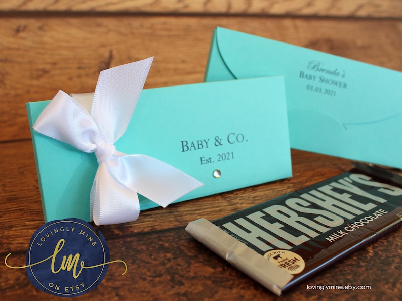 May include: Two rectangular turquoise paper candy bar wrappers with white satin ribbons. One wrapper features the text Baby & Co. Est. 2021 with a small rhinestone, and the other displays Brenda's Baby Shower 03.03.2021. A Hershey's milk chocolate bar is visible.