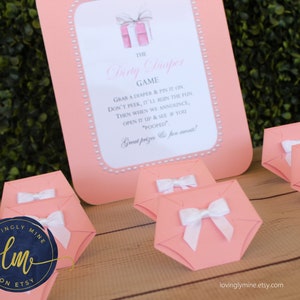 May include: Pink and white diaper-shaped boxes with white bows, used for a baby shower game called "Dirty Diaper Game". The game instructions are printed on a pink sign with white pearl accents. The sign reads "Grab a diaper & pin it on. Don't peek, it'll ruin the fun. Then when we announce, open it up & see if you "pooped". Great prizes & fun await!"