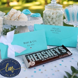 May include: Two rectangular turquoise paper envelopes with white ribbon accents. One envelope features the text "BABY & CO. Est. 2021" and the other "Brenda's BABY SHOWER 03.03.2021". A Hershey's milk chocolate bar is placed in front of the envelopes.