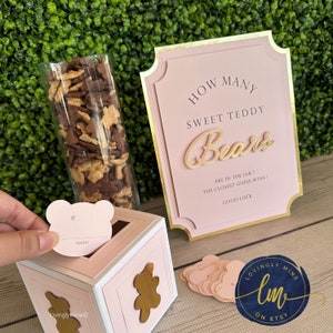 May include: Pink and gold teddy bear themed game for a baby shower. The game asks guests to guess how many teddy bears are in a jar. Guests write their guess on a pink bear-shaped card and drop it into a pink and white box. The box has a gold teddy bear on the front. The sign says "How many sweet teddy bears are in the jar? The closest guess wins! Good luck."