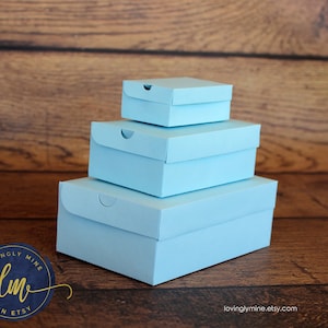 May include: Three light blue gift boxes stacked on top of each other on a wooden surface. The boxes are all the same shape and size, but the top box is slightly smaller than the other two. The boxes are made of a sturdy cardboard material and have a smooth, matte finish.
